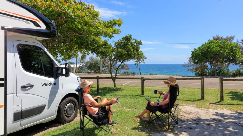 Two women sitting in camp chairs by their caravan with sea views enjoying a cuppa