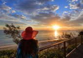 A woman wearing a hat looks out to sea as the sun is setting